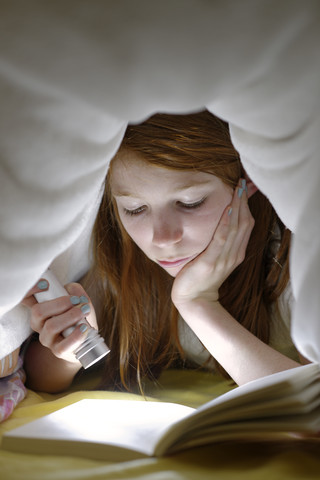 Porträt eines Mädchens, das heimlich im Bett unter der Decke ein Buch liest, lizenzfreies Stockfoto