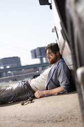 Man lying on ground leaning against car - FMKF001206