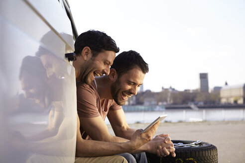 Two laughing men sitting in car looking at digital tablet - FMKF001235