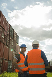 Two men with safety helmets and reflective vests talking at container port - UUF000416