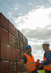 Two men with safety helmets and reflective vests talking at container port - UUF000417