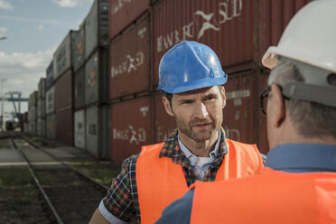 Two men with safety helmets and reflective vests talking at container port - UUF000418