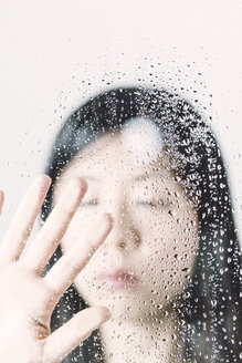 Asian woman with closed eyes behind window pane with water drops - FLF000424