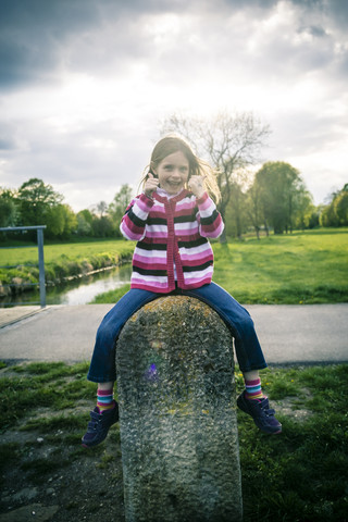 Kleines Mädchen sitzt auf großem Stein mit Daumen hoch, lizenzfreies Stockfoto