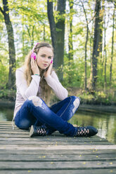 Porträt eines weiblichen Teenagers mit rosa Kopfhörern, der auf einem Holzsteg sitzt - SARF000560