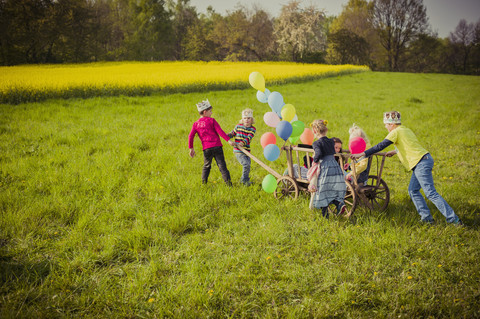 Sechs Kinder unterwegs mit Holzwagen und Luftballons, lizenzfreies Stockfoto