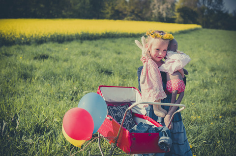 Lächelndes kleines Mädchen mit Puppe und Stofftier auf einer Wiese mit Puppenwagen stehend, lizenzfreies Stockfoto