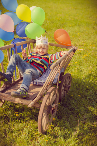 Kleiner Junge liegt auf einem Holzwagen und hält Luftballons, lizenzfreies Stockfoto