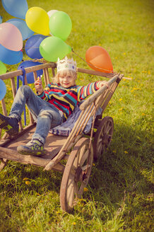 Kleiner Junge liegt auf einem Holzwagen und hält Luftballons - MJF001145