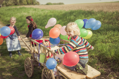 Drei Kinder unterwegs mit Holzwagen und Luftballons, lizenzfreies Stockfoto