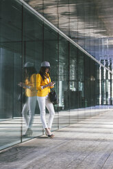 Spanien, Katalonien, Barcelona, junge moderne Frau mit gelber Jacke, die an einer Glasfassade lehnt - EBSF000199