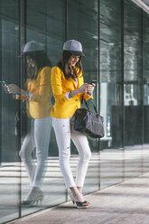 Spanien, Katalonien, Barcelona, junge moderne Frau mit gelber Jacke, die an einer Glasfassade lehnt - EBSF000216