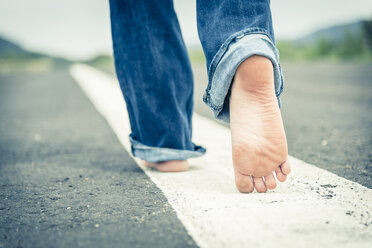 Young woman walking barefoot on centre line of empty street, partial view - ABAF001336