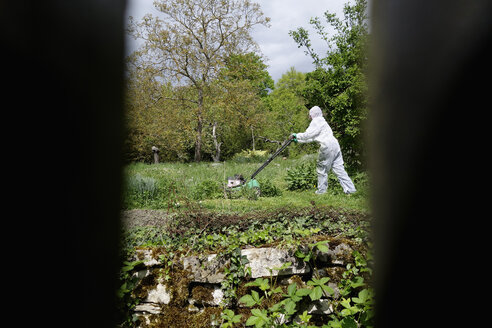 Blick durch Holzgitter auf Frau in weißem Schutzanzug beim Rasenmähen - NDF000443