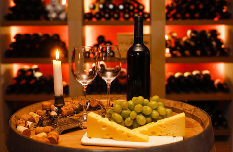 Privater Weinkeller mit einer Flasche Rotwein, zwei Weingläsern, Weintrauben, Käse und einer brennenden Kerze im Vordergrund, lizenzfreies Stockfoto