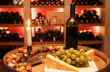 Privater Weinkeller mit einer Flasche Rotwein, zwei Weingläsern, Weintrauben, Käse und einer brennenden Kerze im Vordergrund - JTF000548