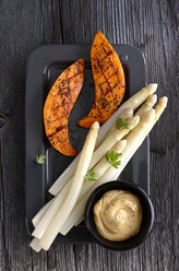 Dish of cooked white asparagus, slices of grilled sweet potato and bowl of dip on grey wood - KSWF001285