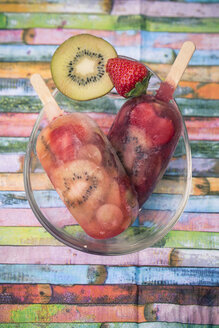 Glass bowl with two kiwi strawberry ice lollies, strawberry and slice of kiwi on coloured wood, elevated view - SARF000654