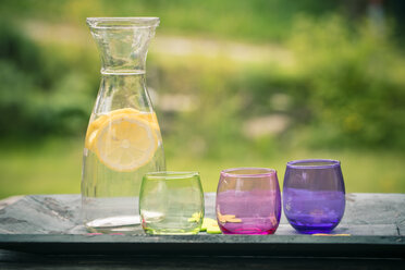 Karaffe Limonade mit Zitronenscheiben und drei farbigen Gläsern auf Holztablett - SARF000655