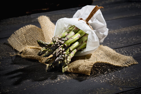 Bündel grüner Spargel in weißer Papiertüte auf Jute und dunklem Holz - MAEF008352