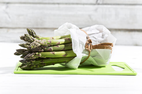 Bündel grüner Spargel in weißer Papiertüte auf grünem Schneidebrett und weißem Holz liegend - MAEF008338