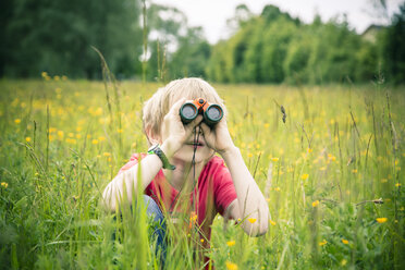 Little boy sitting on meadow watching something with binocular - SARF000658