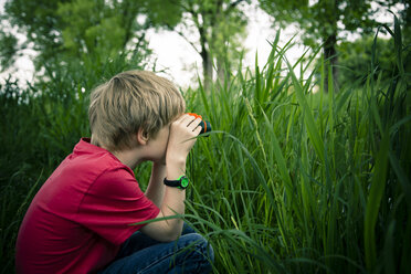 Kleiner Junge sitzt auf einer Wiese und beobachtet etwas mit einem Fernglas - SARF000660