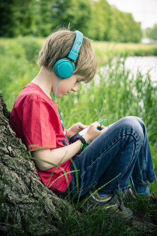Porträt eines Jungen mit Smartphone und Kopfhörern, der an einem Baum lehnt, lizenzfreies Stockfoto