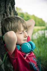Porträt eines entspannten Jungen mit Kopfhörern, der an einem Baum lehnt - SARF000662