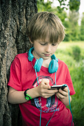 Porträt eines Jungen mit Smartphone und Kopfhörern, der an einem Baum lehnt - SARF000663