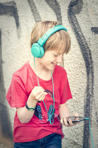 Porträt eines glücklichen Jungen mit Smartphone und Kopfhörern vor einer Fassade, lizenzfreies Stockfoto