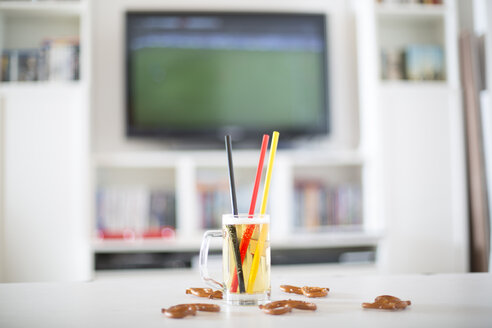 Bierkrug mit drei Trinkhalmen in den deutschen Nationalfarben und Brezeln vor einem Fernseher mit leerem Fußballfeld - SARF000666