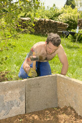 Young man fixing stone slabs for a raised bed with a hammer in the garden - LAF000897