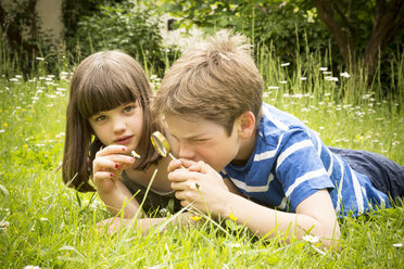 Portrait of brother and sister lying on meadow in the garden having fun with magnifying glass - LVF001375