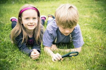 Brother and sister lying on meadow watching flowers with magnifying glass - SARF000673