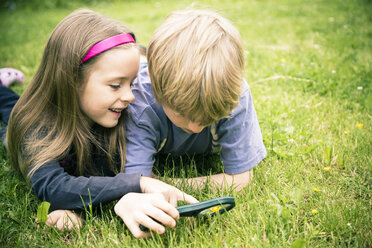 Brother and sister lying on meadow watching flowers with magnifying glass - SARF000675