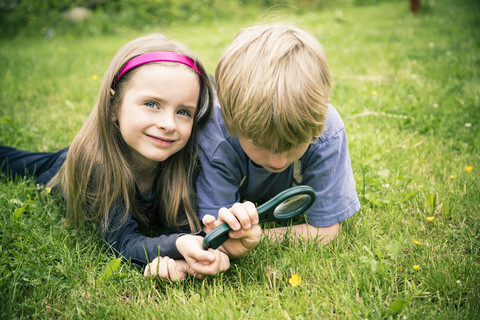 Bruder und Schwester liegen auf einer Wiese und betrachten Blumen mit einem Vergrößerungsglas, lizenzfreies Stockfoto