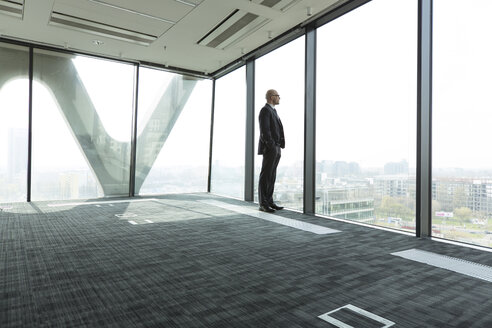 Businessman on empty office floor looking out of window - WESTF019467