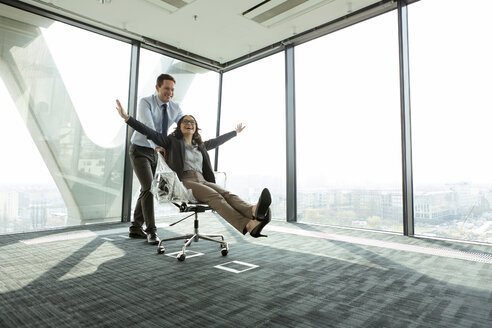 Businessman pushing businesswoman in office chair - WESTF019494