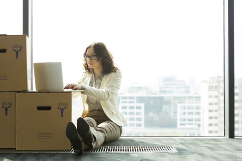 Businesswoman using laptop on empty office floor with cardboard boxes - WESTF019500