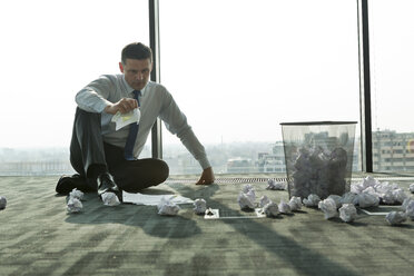 Businessman sitting on office floor surrounded by crumpled paper - WESTF019298