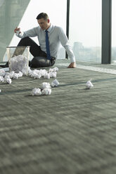 Businessman sitting on office floor surrounded by crumpled paper - WESTF019300