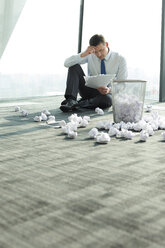 Businessman sitting on office floor surrounded by crumpled paper - WESTF019425