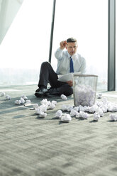 Businessman sitting on office floor surrounded by crumpled paper - WESTF019406