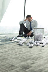 Businessman sitting on office floor surrounded by crumpled paper - WESTF019407