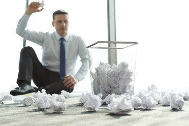 Businessman sitting on office floor surrounded by crumpled paper - WESTF019408