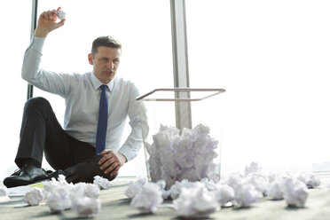 Businessman sitting on office floor surrounded by crumpled paper - WESTF019426