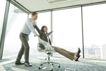 Businessman pushing businesswoman in office chair - WESTF019402