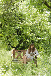 Lächelnde junge Frau mit einem Buch auf einer Holzbank in einem Garten - LAF000891
