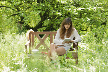 Junge Frau liest ein Buch auf einer Holzbank in einem Garten - LAF000892